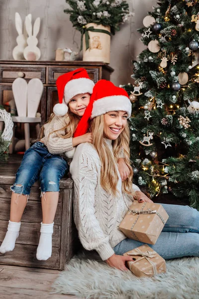 산타 클라우스 모자를 쓴 행복 한 여인이 크리스마스 트리 근처에 딸과 앉아 있는 모습 — 스톡 사진