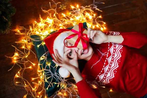Divertida linda mujer tumbada en el suelo y sosteniendo presente en la habitación con decoraciones de Navidad en casa — Foto de Stock
