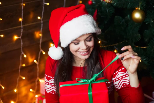 Feliz joven en Santa Claus sombrero regalo de apertura en casa cerca del árbol de Navidad. — Foto de Stock