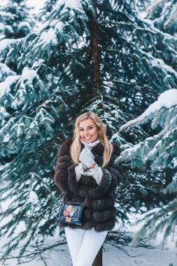 Kürk giymiş gülümseyen kadın kışın karlı ormanda yürüyor..