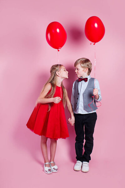 Valentines day. Little boy kissing beautiful little girl on pink background. Children holding heart balloons