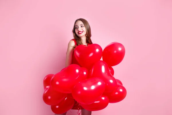 Sevgililer Günü. Pembe arka planda kalp şeklinde kırmızı balonları olan kırmızı elbiseli güzel, zarif genç bir kadın.. — Stok fotoğraf
