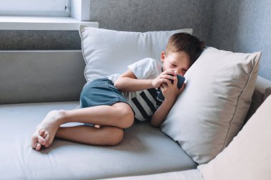 Küçük çocuk kanepede uzanıyor ve evde telefonla oynuyor.