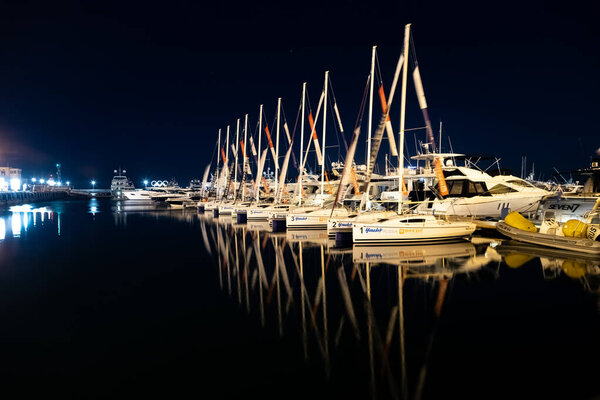 Маленькие спортивные яхты для парусной регаты на пирсе в Сочи стоят ночью в ряд в свете фонарей, красиво отраженных в тихой воде.