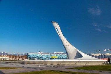 The Big Olympic Torch in Sochi against the backdrop of mountains with the flags of the countries and the Iceberg Ice Palace on a sunny day.
