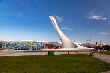 The Big Olympic Torch in Sochi against the backdrop of mountains with the flags of the countries and the Iceberg Ice Palace on a sunny day.