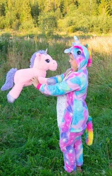 Kigurumi 'de bir çocuk. Gökkuşağı tek boynuzlu at kostümlü bir kız sıcak bir yaz akşamında dışarıda duruyor..