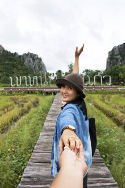 Thai woman lead man by the hand and hold on walkway wooden bridge in rice field of landmarks KaoNor KaoKaew Limestone mountains at Banphot Phisai city on October 19,2020 in Nakhon Sawan, Thailand clipart