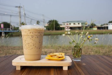 Latte or mocha or cappuccino iced coffee serve with bread cookie and grass flower on wooden table for travelers thai people rest relax eat and drink beside irrigation canal at Nonthaburi, Thailand