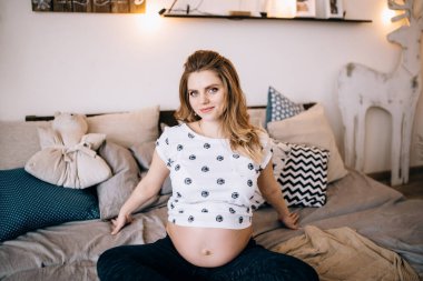 Hamilelik, dinlenme, insanlar ve beklenti konsepti. Mutlu gülümseyen hamile bir kadının evde oturup karnına dokunması.