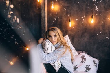 Güzel sarışın yastıkla yatakta oturuyor.