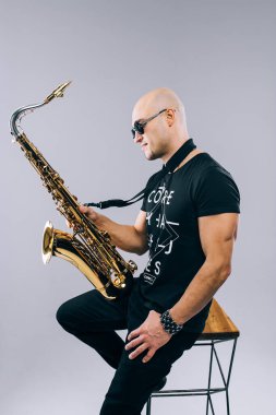 Güneş gözlüklü ve saksafonlu bir adam stüdyoda, gri arka planda.