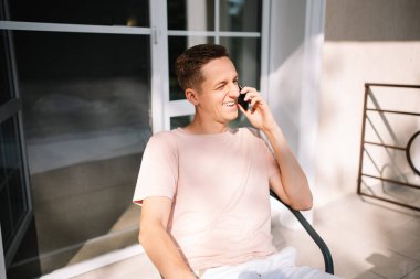 Balkonda telefonda konuşan bir adam sandalyede oturuyor.