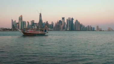 Doha ufuk çizgisinden gün batımına doğru zumluyor Arap körfezinde Katar bayrakları ve arka planda bulutlar görünüyor.