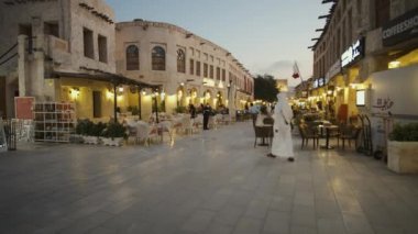 Souq Waqif, Doha Katar ana caddesinde, geceleri yakınlaşırken, arka planda Katar bayrağı ile oturan ve yürüyen yerli ve ziyaretçileri gösteriyor.