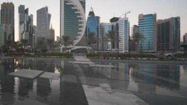 Doha Katar 'daki batı körfezi gökdelenleri öğleden sonra Sheraton parkından çekilmiş ön planda fıskiyeler, Katar bayrakları, palmiye ağaçları ve Corniche caddesinde hareket eden arabalar.