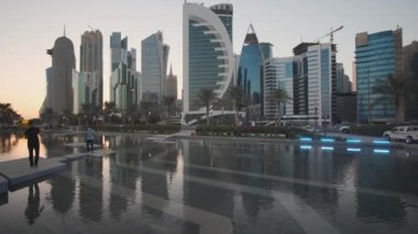 Doha Katar 'daki batı körfezi gökdelenleri, Sheraton parkından ön planda fıskiyeler, Katar bayrakları, palmiye ağaçları ve arabalarla Corniche caddesinde çekilen görüntüler.