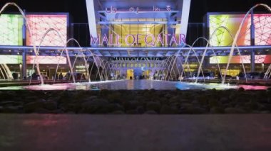 Gece Doha Katar 'daki Katar AVM' si ön plandaki aydınlatma çeşmesini ve arka plandaki ana girişi, hareket halindeki insan ve arabaları gösteriyor.