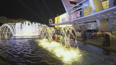 Doha Katar 'daki Katar AVM' si geceleri yakınlaşıyor ve ön planda yanan çeşme ve arka planda insanlar ve arabaların hareket ettiği ana giriş kapısını gösteriyor.
