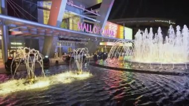 Doha Katar 'daki Katar AVM' si geceleri yakınlaşıyor ve ön planda yanan çeşme ve arka planda insanlar ve arabaların hareket ettiği ana giriş kapısını gösteriyor.