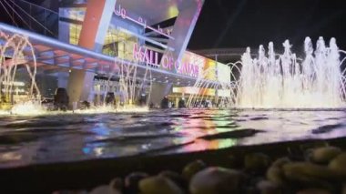 Doha Katar 'daki Katar AVM' si geceleri yakınlaşıyor ve ön planda yanan çeşme ve arka planda insanlar ve arabaların hareket ettiği ana giriş kapısını gösteriyor.