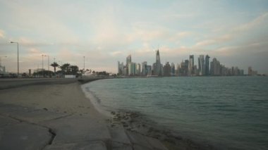 Doha ufuk çizgisi Corniche gezinti alanından öğleden sonra yakınlaştırıyor. Arap körfezinde Katar bayrakları ve arka planda bulutlar görünüyor.