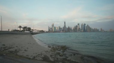Doha ufuk çizgisi Corniche gezinti alanından öğleden sonra yakınlaştırıyor. Arap körfezinde Katar bayrakları ve arka planda bulutlar görünüyor.