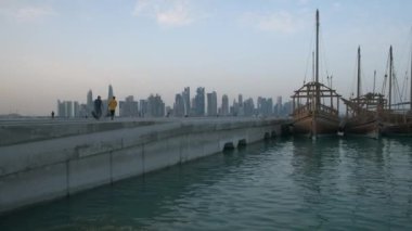Doha ufuk çizgisinden gün batımına doğru zumluyor Arap körfezinde Katar bayrakları ve arka planda bulutlar görünüyor.