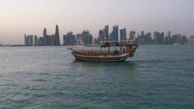 Doha ufuk çizgisinden gün batımına doğru zumluyor Arap körfezinde Katar bayrakları ve arka planda bulutlar görünüyor.