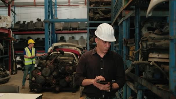 白人业主和亚裔工人 两个人在仓库里检查发动机零件 他们两个人都在工作时戴着钢盔 所以大部分产品都是汽车行业的机器 — 图库视频影像