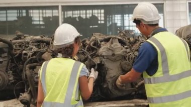 Üniformalı ve kasklı Asyalı erkek ve kadın işçiler, yedek araba deposu fabrikasının personel stok kontrolü. Otomotiv endüstrisi için eski motorlardan ve makine tekniklerinden bahsediyorlar..