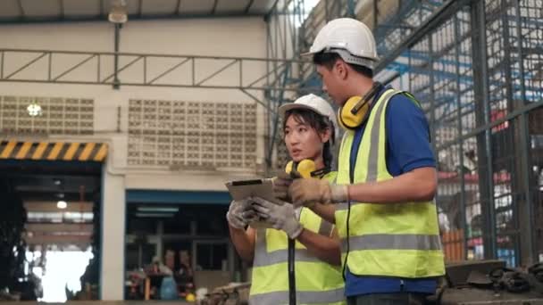 身穿制服头戴钢盔的亚裔男女 两个工人都在工业工厂 使用平板电脑对仓库中的货物和产品进行检查 向同一家工厂的同事解释 — 图库视频影像