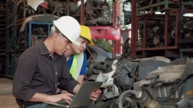 Kafkas Sahibi & Asyalı İşçi: Bir depoda motor parçalarını kontrol eden iki adam. Her ikisi de iş sırasında kask takıyor. Ürünlerin çoğu otomobil endüstrisindeki makineler..