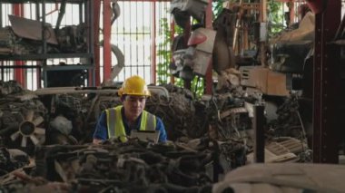 Güvenlik üniforması ve miğferi olan genç Asyalı erkek işçi. Bir depo fabrikasındaki otomobil endüstrisindeki motor ve makineler gibi yedek parça stoklarını kontrol etmek için bir tablet kullanmak.