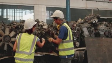 Üniformalı ve kasklı Asyalı erkek ve kadın işçiler, yedek araba deposu fabrikasının personel stok kontrolü. Otomotiv endüstrisi için eski motorlardan ve makine tekniklerinden bahsediyorlar..