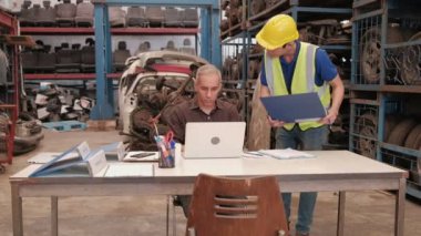 Otomotiv deposunda iki adam yedek depoda: Kafkas sahibi siparişleri kontrol etmek için dizüstü bilgisayar kullanırken, Asyalı erkek işçiler otomobil endüstrisinde yığılmış çok sayıda yedek motor parçalarını inceliyor.