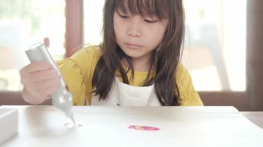 Sarı tişörtlü ve önlüklü şirin Asyalı kız evde beyaz kağıda çok renkli simli boya çizen tek el yazısı. Sanat dersi çocukları yaratıcı yapar. Okul becerilerini geliştirmek için eğlenceli aktiviteler.