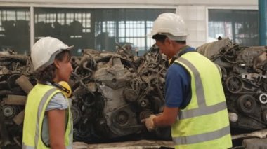 Üniformalı ve kasklı Asyalı erkek ve kadın işçiler, yedek araba deposu fabrikasının personel stok kontrolü. Otomotiv endüstrisi için eski motorlardan ve makine tekniklerinden bahsediyorlar..