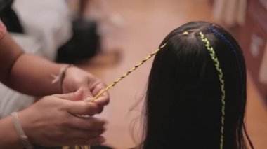 Yakın plan VDO: Güzel, uzun siyah saçlı Asyalı kız güzellik salonunda çok renkli ipliklerle örülüyor. Çocuk modası, örgü ören örgü ören örgü ören yün örgüler..