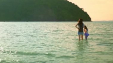 Bulanık Odaklı VDO: Bir anne akşam vakti sevgiyle kızının elini denizde yürüyüşe çıkardı. Yazın çocukların tatili sırasında plajda turuncu ışıklar ve gün batımları varken.