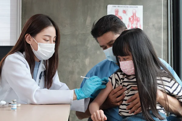 Female doctor with face mask vaccinating Asian girl to prevent coronavirus (COVID) 19) At the pediatrics clinic in kid hospital with father nearby. Injections treat illnesses, cause pain in children.