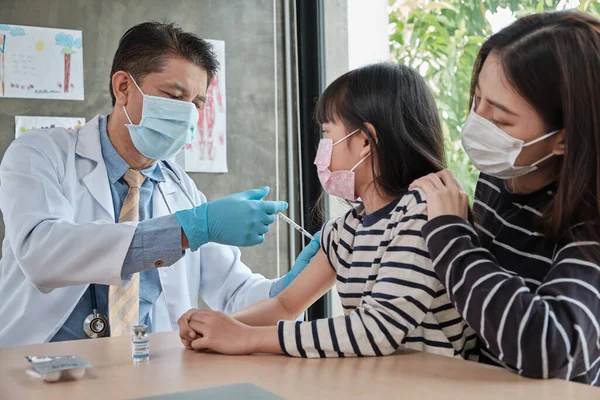 Male doctor with face mask vaccinating Asian girl to prevent coronavirus (COVID) 19) At the pediatrics clinic in kid hospital with mother nearby. Injections treat illnesses, cause pain in children.