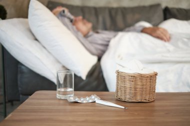 Asyalı adam işe gitmemiş ve hastalık nedeniyle hastalık izni almış, termometreyle ateşi ölçmüş, ilaç almış, evdeki kanepede battaniyelerle uyumuş, peçeteler ve kapsül hapları masada..