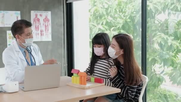 顔のマスクをした母親は 病気の娘を子供病院の小児科のアジア系男性医師との試験予約に連れて行きます 病気を治療し 家族の健康を相談する — ストック動画
