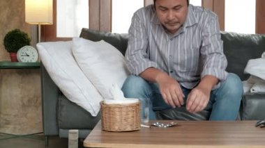 Asyalı ofis çalışanı hastalık nedeniyle izinde, termometre ateşini ölçüyor, ilaç alıyor, evdeki kanepede battaniyeler içinde uyuyor, mendiller ve haplar masada..