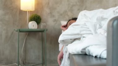 Asyalı ofis çalışanı işe gitmemiş, hastalık yüzünden geç kalkmış ve hastalık izni almış, ilaçlarını almış, evdeki kanepede beyaz battaniyelerle uyumuş, mendiller ve gri beton duvarın önündeki masanın üzerinde haplar varmış..