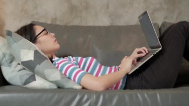 Günlük serbest çalışan Asyalı kadın e-ticaret işinde çalışıyor ve internet üzerinden öğrenim görüyor, dizüstü bilgisayarını kullanıyor oturma odasındaki kanepede uzanıyor ve sohbet ediyor, yeni normal yaşam tarzı....