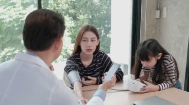 Asyalı erkek doktor sağlıklı danışmanlık, klinikte anne olan bir kadının tansiyon kontrolü. İlaç tedavisi için akıllı telefondan işlem görüyor, bir çocuk, kızı yakınlarda..