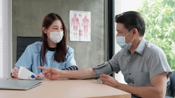 顔のマスクをしたアジア系女性医師が病院の血圧モニターで車椅子高齢者の健康診断を行う トークセラピーは看護学の試験です — ストック動画