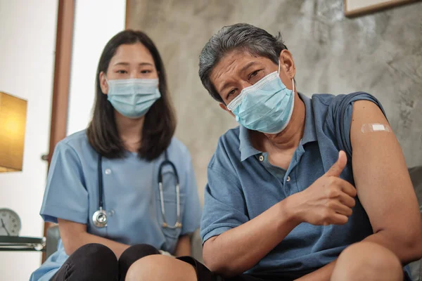 Elderly patient man with face mask thumb up when vaccinated by Asian female, prevent illness from virus infection. Home healthcare by professionals to treat sick people who are retired.